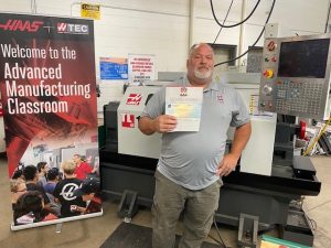 dan leatzow poses in front of a machine holding a certificate