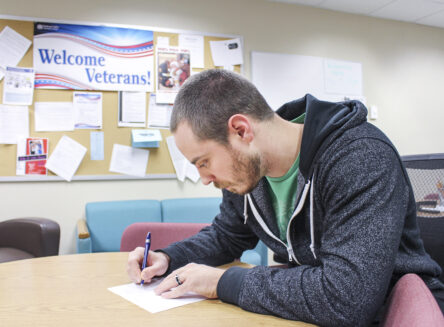 a student writes at a table with a sign reading veterans center in the background
