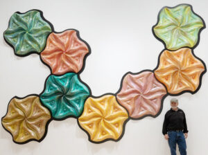 sculpture artist Chip Clawson posing in front his large artwork, taking up the space of an entire wall behind him