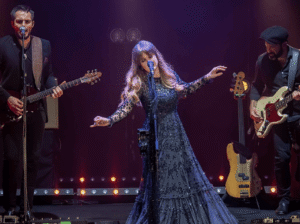 The Rumours of Fleetwood Mac tribute band performs on stage. A female singer in a long, dark gown stands center stage, holding a microphone and gesturing with her arms. A male guitarist and a male bassist are on either side of her, playing their instruments.