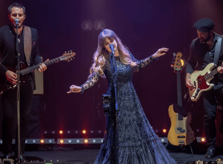 The Rumours of Fleetwood Mac tribute band performs on stage. A female singer in a long, dark gown stands center stage, holding a microphone and gesturing with her arms. A male guitarist and a male bassist are on either side of her, playing their instruments.