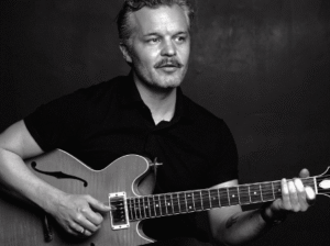 A black-and-white photo of folk artist Kristian Matsson, also known as The Tallest Man on Earth. He is seated, looking to the side, and playing a hollow-body electric guitar.