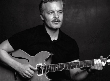 A black-and-white photo of folk artist Kristian Matsson, also known as The Tallest Man on Earth. He is seated, looking to the side, and playing a hollow-body electric guitar.