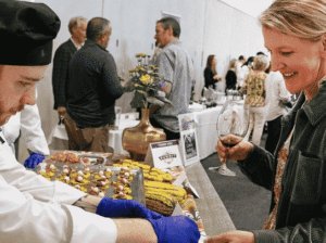 An FVCC Culinary Institute student, wearing a black chef's hat and blue gloves, serves a dish to a smiling woman holding a glass of wine. The event is set up with tables of food and other guests in the background.