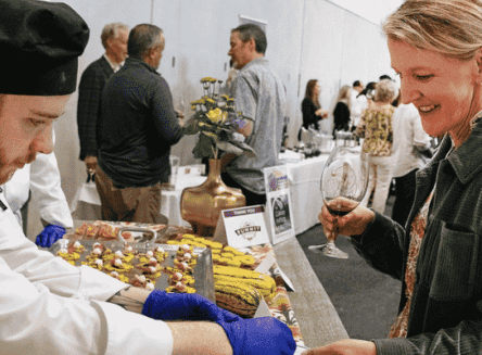An FVCC Culinary Institute student, wearing a black chef's hat and blue gloves, serves a dish to a smiling woman holding a glass of wine. The event is set up with tables of food and other guests in the background.