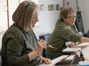Two women, students in an FVCC Senior Institute creative arts class, are seated at a table. The woman in the foreground is holding a paintbrush and looking down at her artwork.