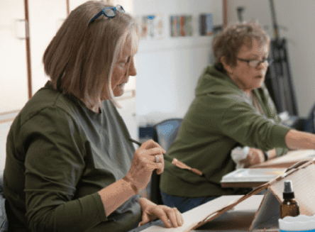 Two women, students in an FVCC Senior Institute creative arts class, are seated at a table. The woman in the foreground is holding a paintbrush and looking down at her artwork.