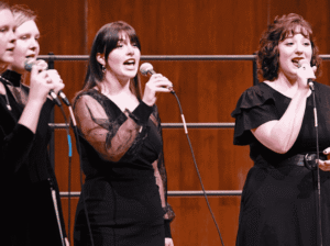 A group of four female singers from the FVCC vocal ensemble, dressed in black, perform on stage with microphones.