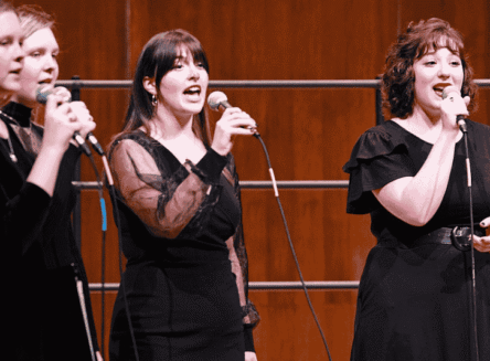 A group of four female singers from the FVCC vocal ensemble, dressed in black, perform on stage with microphones.