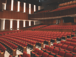 A wide shot of the Wachholz College Center's McLaren Hall, showing rows of red upholstered seats and the stage from the audience's perspective. The room is well-lit, with wooden panels and balcony seating visible.
