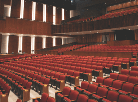 A wide shot of the Wachholz College Center's McLaren Hall, showing rows of red upholstered seats and the stage from the audience's perspective. The room is well-lit, with wooden panels and balcony seating visible.