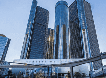 A low-angle shot of the modern glass skyscrapers of the General Motors Renaissance Center against a clear blue sky. A walkway is visible in the foreground.