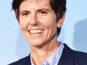 A headshot of comedian Tig Notaro smiling, wearing a light blue button-down shirt and a dark blazer.