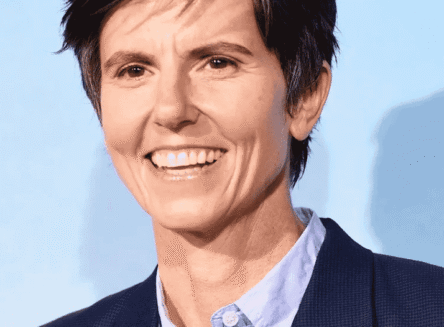 A headshot of comedian Tig Notaro smiling, wearing a light blue button-down shirt and a dark blazer.