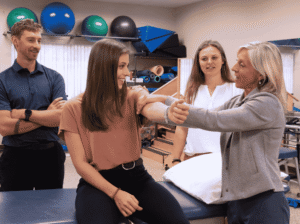 A female instructor demonstrates a physical therapy technique on a student's arm while two other students observe in a well-equipped classroom featuring treatment tables and exercise balls.