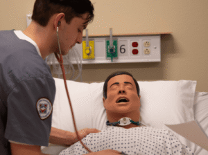Flathead Valley Community College student in gray scrubs uses a stethoscope to listen to the chest of a medical simulation mannequin lying in a hospital bed.