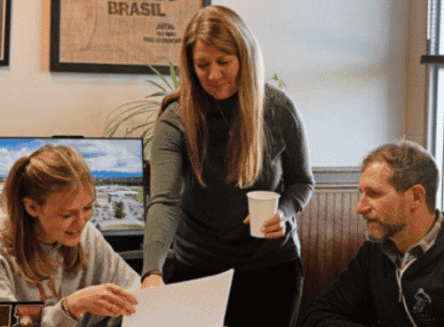 Two faculty members and one student sit around a wooden table in an office, reviewing documents together.