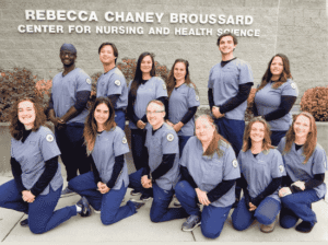 Twelve nursing graduates in blue scrubs pose together in front of the Rebecca Chaney Broussard Center for Nursing and Health Science building.
