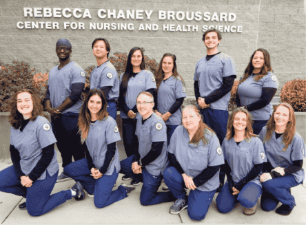 Twelve nursing graduates in blue scrubs pose together in front of the Rebecca Chaney Broussard Center for Nursing and Health Science building.