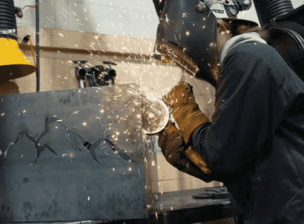 A person wearing a black welding helmet and protective brown gloves uses a grinder on a metal sheet, creating a spray of sparks in a workshop.