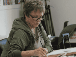 A smiling woman with short grey hair and glasses painting a landscape on a canvas with a paintbrush.