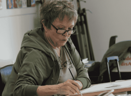 A smiling woman with short grey hair and glasses painting a landscape on a canvas with a paintbrush.