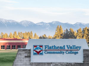 The Flathead Valley Community College stone entrance sign with the blue and red mountain logo, set against a backdrop of snow-capped mountain range.
