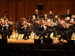 A large group of violinists and a harpist perform on a stage with warm wooden paneling during a Glacier Symphony concert.