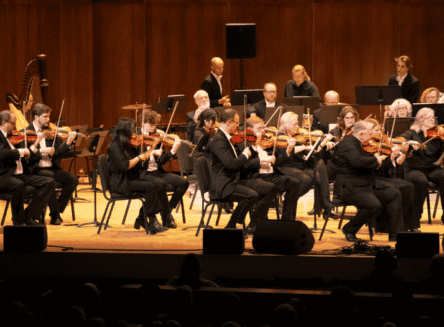 A large group of violinists and a harpist perform on a stage with warm wooden paneling during a Glacier Symphony concert.