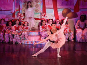 A male and female ballet dancer perform a graceful pose on a festive stage decorated with candy canes, while child dancers in red and white striped costumes sit in the background.