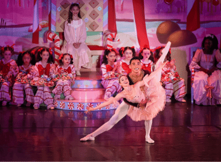 A male and female ballet dancer perform a graceful pose on a festive stage decorated with candy canes, while child dancers in red and white striped costumes sit in the background.