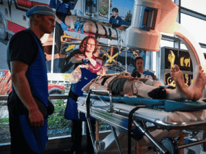 A group of medical faculty and students in blue scrubs performing a medical simulation on a mannequin in a hospital bed at the Broussard at the Rebecca Chaney Broussard Center for Nursing & Health Science.