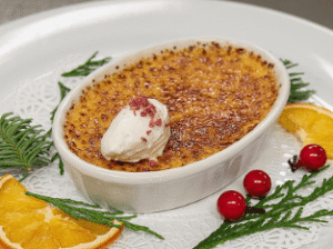 A close-up photo of a festive holiday dessert, likely crème brûlée, served in an oval white ramekin with a perfectly caramelized sugar crust. The dish is topped with a dollop of whipped cream and red crumbles. It sits on a white plate with a decorative lace doily, garnished with orange slices, evergreen sprigs, and bright red berries.