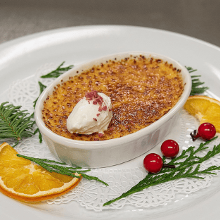 A close-up photo of a festive holiday dessert, likely crème brûlée, served in an oval white ramekin with a perfectly caramelized sugar crust. The dish is topped with a dollop of whipped cream and red crumbles. It sits on a white plate with a decorative lace doily, garnished with orange slices, evergreen sprigs, and bright red berries.