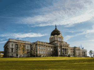 A wide-angle shot of the Montana State Capitol building in Helena under a blue sky with light clouds. The building's iconic copper dome is centered, surrounded by green lawns.