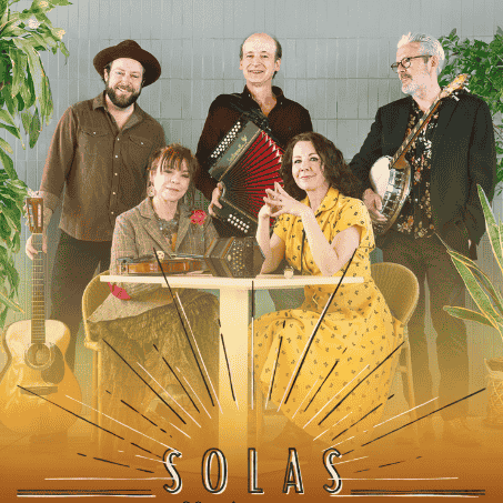 Five members of the acclaimed Irish traditional music group Solas are posed together in a bright, modern setting surrounded by potted plants. Two women are seated at a white table, and three men are standing behind them, holding traditional instruments including a guitar, an accordion (played by original member John Williams), and a banjo.