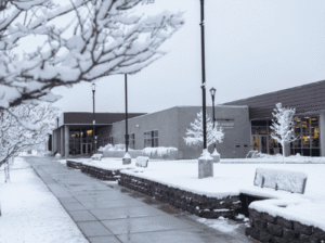 A serene, snow-covered view of the exterior of the Broussard Family Library and Learning Commons at Flathead Valley Community College. A wet, cleared sidewalk leads toward the modern building, flanked by lamp posts and trees heavily blanketed in fresh snow under an overcast winter sky.