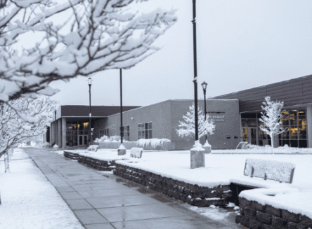A serene, snow-covered view of the exterior of the Broussard Family Library and Learning Commons at Flathead Valley Community College. A wet, cleared sidewalk leads toward the modern building, flanked by lamp posts and trees heavily blanketed in fresh snow under an overcast winter sky.