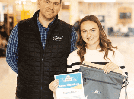 A young woman holding an award certificate and a blue Nike drawstring bag stands next to a man wearing a Flathead Insurance vest.