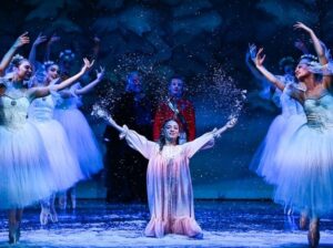 A group of ballet dancers in white tutus and a central dancer in a pink gown perform on stage while artificial snow falls.