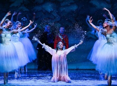 A group of ballet dancers in white tutus and a central dancer in a pink gown perform on stage while artificial snow falls.