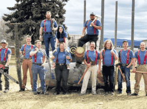 A group of eleven FVCC Logger Sports team members in blue shirts and red suspenders posing outdoors with traditional logging equipment, including crosscut saws, axes, and a chainsaw.