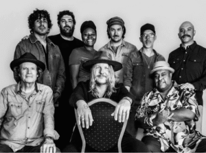 A professional black and white group portrait of nine musicians from the Devon Allman Project and Donavon Frankenreiter, featuring three seated members in the front and six standing behind them.