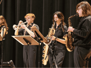 Four FVCC student musicians in black attire performing on stage, including a trumpet player, two saxophone players, and a violinist in the background.