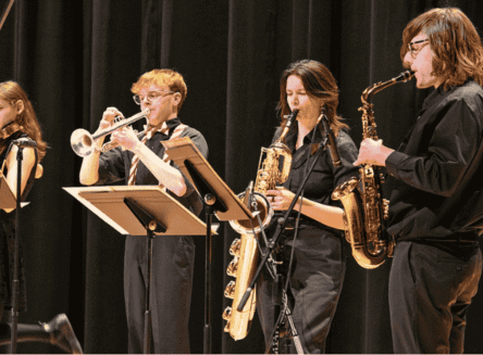 Four FVCC student musicians in black attire performing on stage, including a trumpet player, two saxophone players, and a violinist in the background.