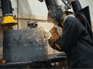 FVCC student in protective welding gear, including a hood and gloves, uses a torch to weld an intricate mountain design onto a metal plate, with bright orange sparks flying.