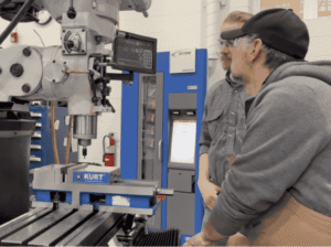 Two individuals standing beside an industrial metal lathe or vertical mill in the FVCC Trades Institute machine shop, surrounded by various machining tools.
