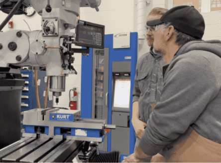 Two individuals standing beside an industrial metal lathe or vertical mill in the FVCC Trades Institute machine shop, surrounded by various machining tools.