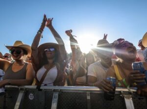 A cheering crowd of people in cowboy hats and sunglasses at an outdoor concert, with the sun shining brightly behind them.