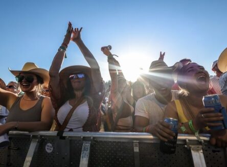 A cheering crowd of people in cowboy hats and sunglasses at an outdoor concert, with the sun shining brightly behind them.